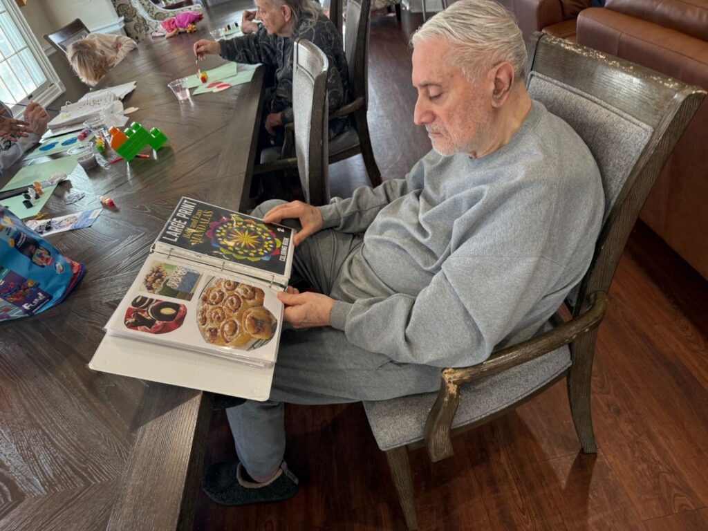 A Fox Trail memory care resident turning the pages of a large print picture book at the dining table while other residents work on crafts